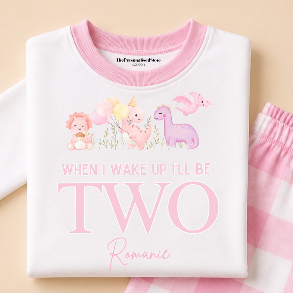 A folded white long-sleeve pajama top with pink trim and a pastel animal graphic, paired with pink-and-white gingham pajama pants on a warm beige background. Visible text includes 'ThePersonalisedStore' branding and the phrase 'When I wake up I'll be two romantic.