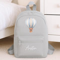 A light gray backpack with a hot air balloon graphic and the name 'Austin' in white script on the front pocket, shown standing on a carpeted floor next to a bed and dresser.