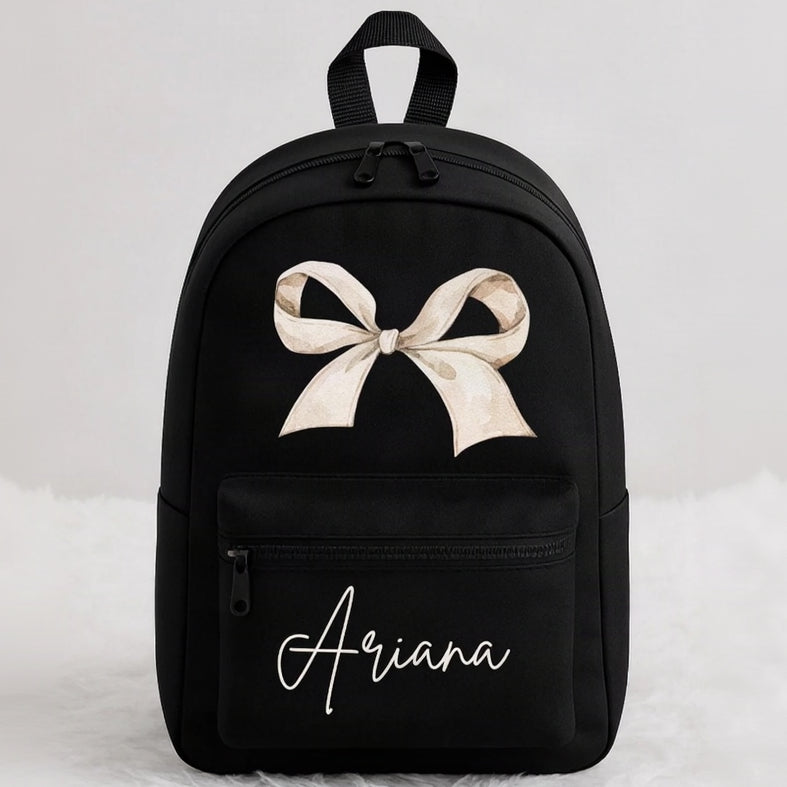 A black backpack with a large beige bow graphic on the front, the name 'Ariana' in white script, and a top handle, shown against a light gray background.