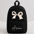 A black backpack with a large beige bow graphic on the front, the name 'Ariana' in white script, and a top handle, shown against a light gray background.