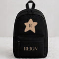 A black backpack with a front zippered pocket, featuring a tan star graphic with the letter 'R' on the upper front and 'REIGN' printed in tan on the lower pocket, set against a light gray background on a white faux fur surface.
