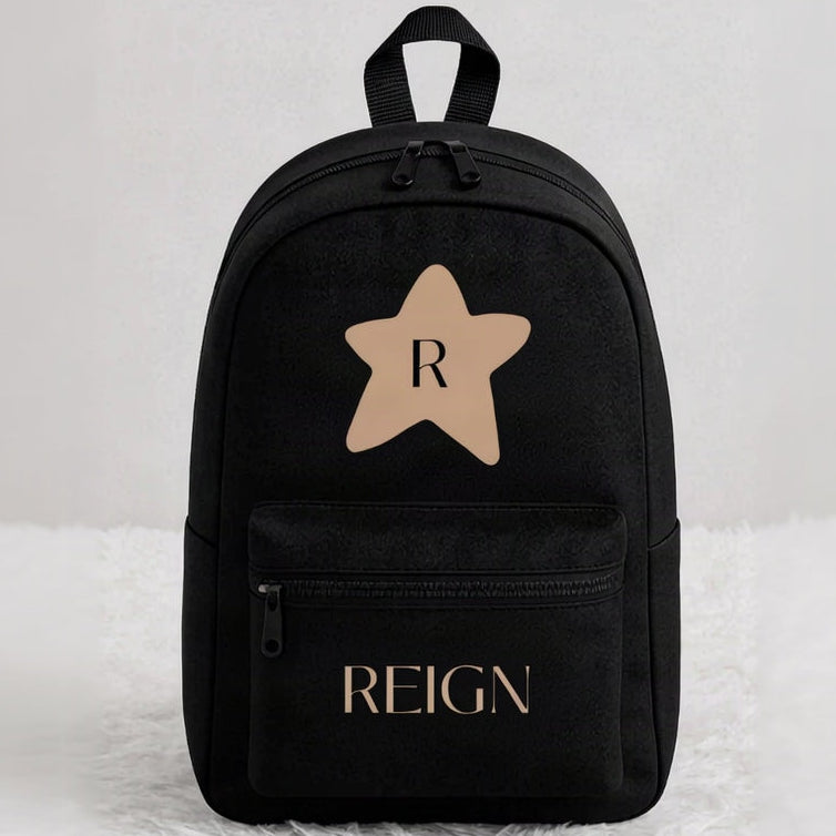 A black backpack with a front zippered pocket, featuring a tan star graphic with the letter 'R' on the upper front and 'REIGN' printed in tan on the lower pocket, set against a light gray background on a white faux fur surface.