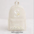 A cream-colored canvas backpack featuring a large white monogram 'S' decorated with daisies, the name 'SAGE' in white text on the front pocket, and a row of small white daisy illustrations at the bottom. The image is set against a clean white background with a watermark at the bottom.