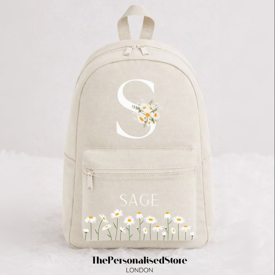 A cream-colored canvas backpack featuring a large white monogram 'S' decorated with daisies, the name 'SAGE' in white text on the front pocket, and a row of small white daisy illustrations at the bottom. The image is set against a clean white background with a watermark at the bottom.
