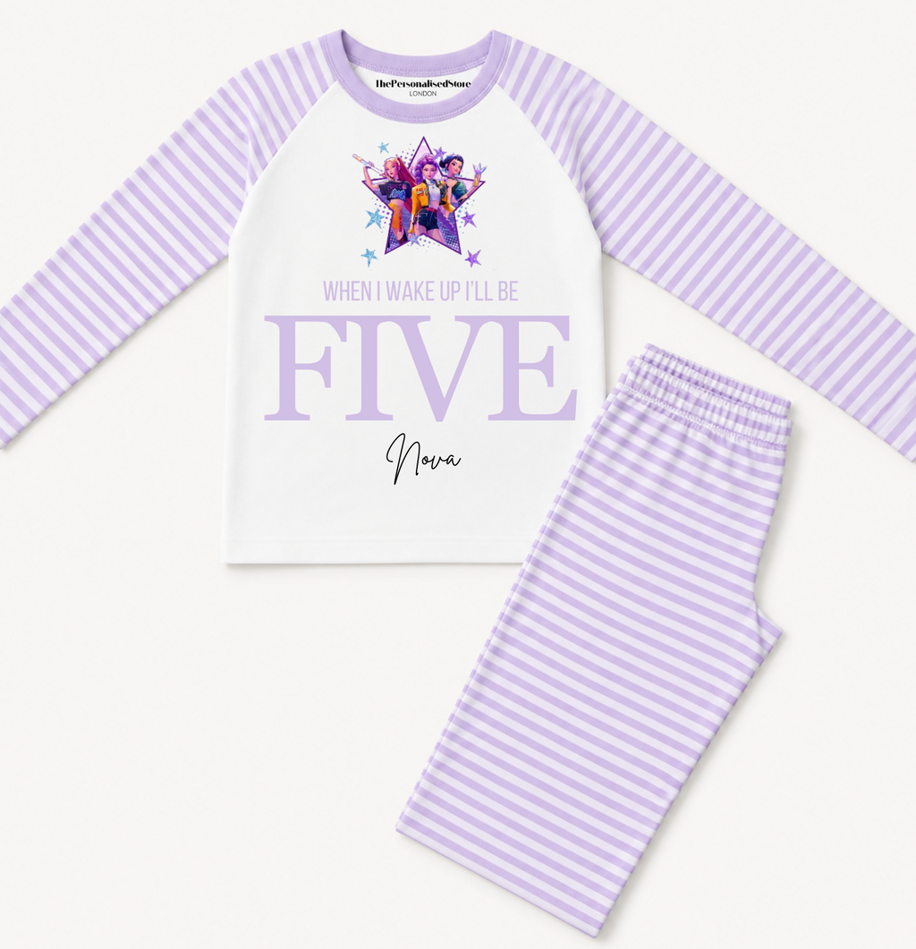 A children's pajama set featuring a long-sleeve top and matching pants. The top is white with light purple stripes on the sleeves and collar, decorated with a colorful graphic and the text 'WHEN I WAKE UP I'LL BE FIVE'.