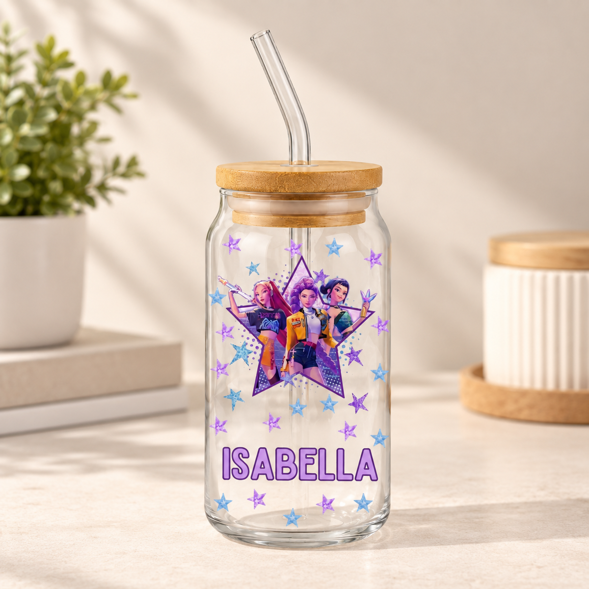 A clear glass tumbler with a bamboo lid and curved glass straw, featuring a colorful star graphic with cartoon characters and the name 'ISABELLA' in purple, set on a light tabletop with a blurred plant and container in the background.
