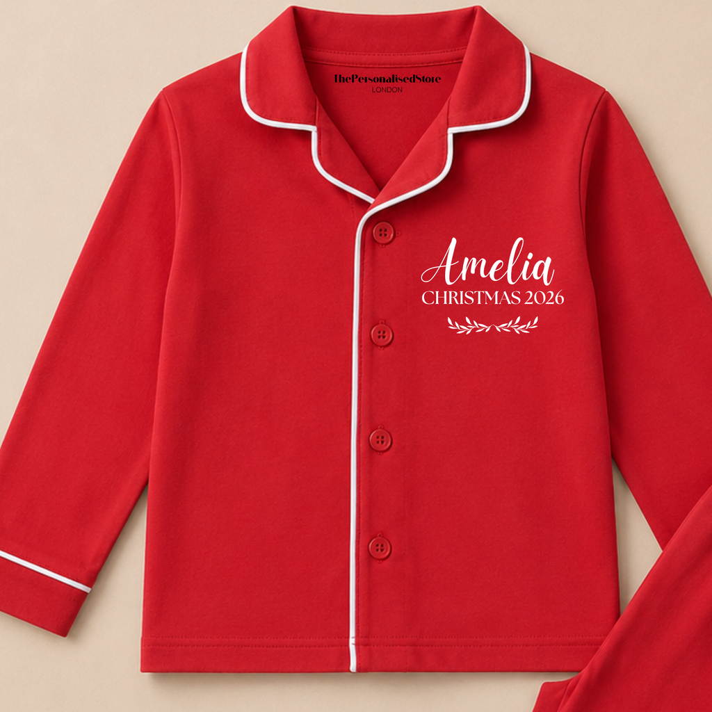 A red two-piece pajama set with white piping trim, featuring a long-sleeve button-front top and matching pants. The top is personalized with the name 'Amelia' and the text 'CHRISTMAS 2026' on the right chest.
