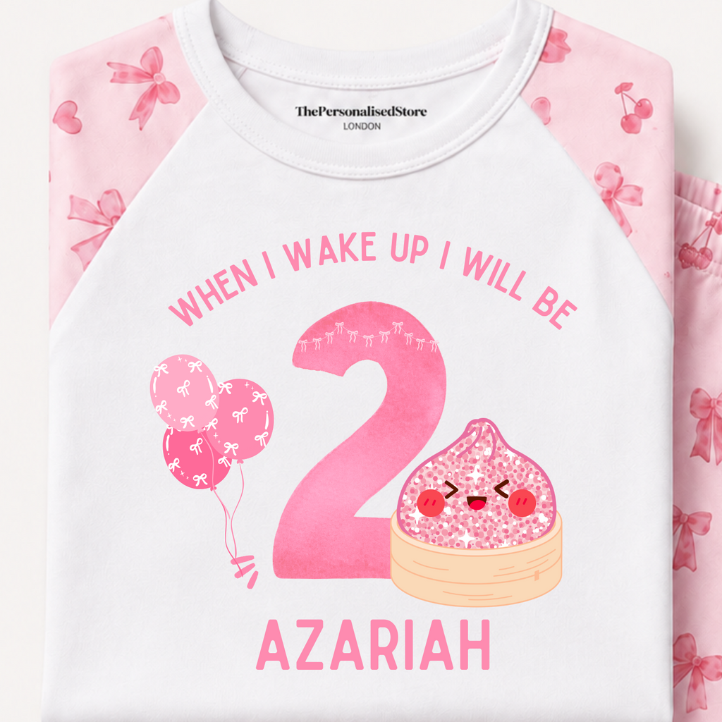 A folded two-piece kids' pajama set on a light cream background. The top is white with pink raglan sleeves and a pink birthday graphic reading 'WHEN I WAKE UP I WILL BE 2 AZARIAH' with balloons and a cake.