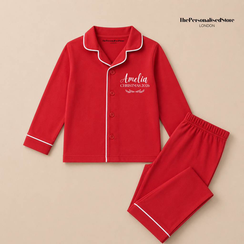 A red two-piece pyjama set with white piping trim, featuring a long-sleeve button-front top and matching pants. The top is personalized with the name 'Amelia' and the text 'CHRISTMAS 2026' and on the right chest.