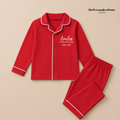 A red two-piece pyjama set with white piping trim, featuring a long-sleeve button-front top and matching pants. The top is personalized with the name 'Amelia' and the text 'CHRISTMAS 2026' and on the right chest.
