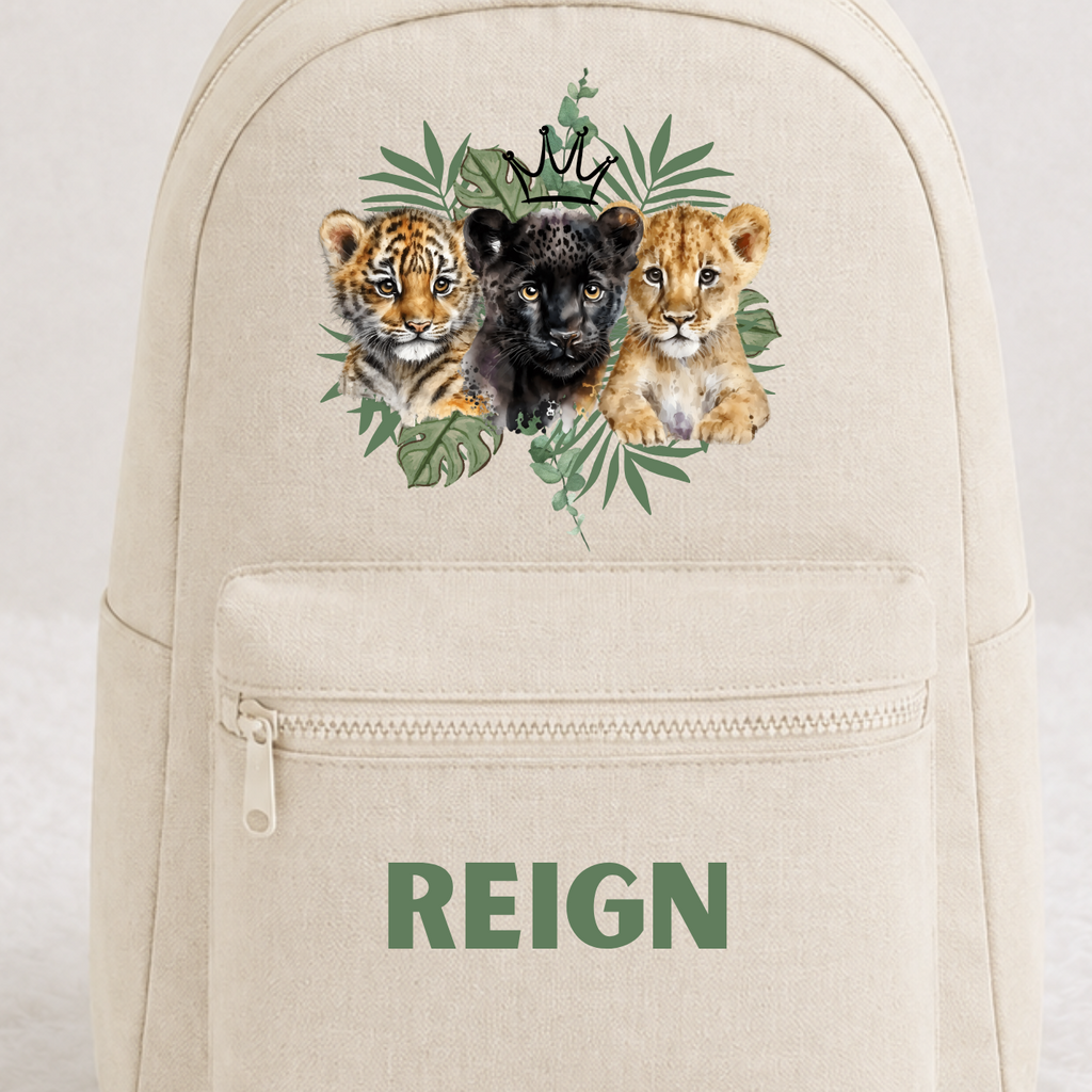 A cream-colored canvas backpack with a front zip pocket, featuring a graphic of a black panther surrounded by green leaves and three lion cubs, with the word 'REIGN' printed in green below. The backpack is displayed on a white faux fur surface against a light gray background.