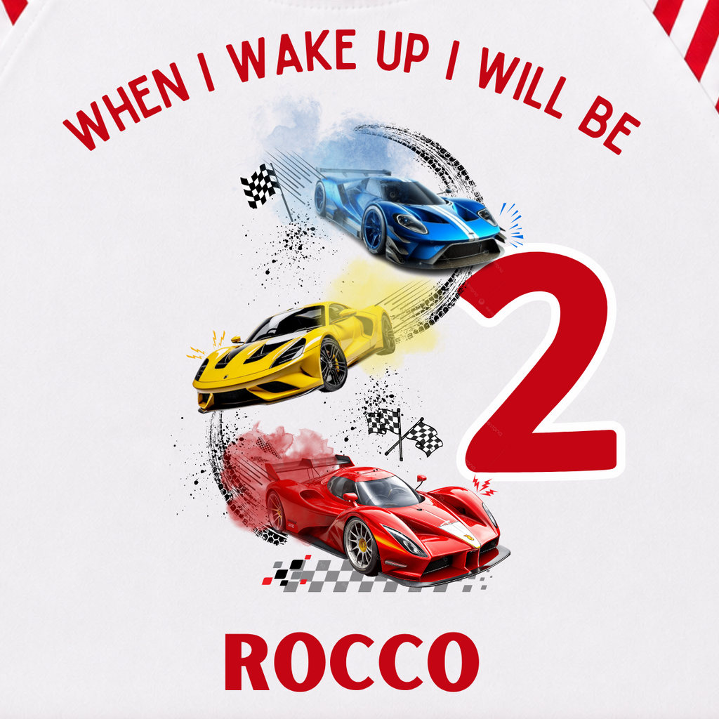 A children's two-piece pajama set laid flat on a light cream background. The long-sleeve top is white with red raglan sleeves and collar, featuring a colorful race car graphic and the text 'WHEN I WAKE UP I WILL BE 2 ROCCO'.